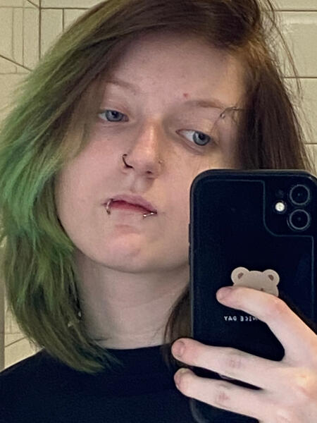 A photo of the host, he’s standing in the walmart bathroom. Glitch has black and green hair, facial piercings and blue eyes.